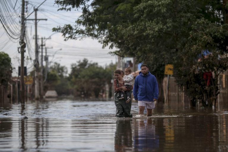 Foto colorida de mulher com criança no colo andando por rua alagada em Porto Alegre. Ao lado delas, aparece um homem também andando em meio às águas. Desastre mostram relação entre saúde materna e mudanças climáticas