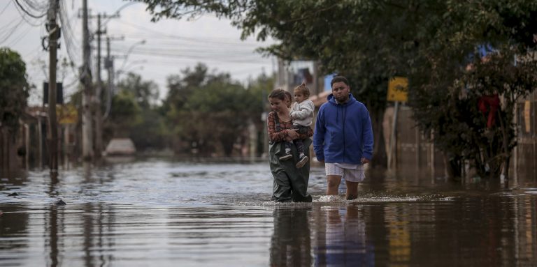 Foto colorida de mulher com criança no colo andando por rua alagada em Porto Alegre. Ao lado delas, aparece um homem também andando em meio às águas. Desastre mostram relação entre saúde materna e mudanças climáticas