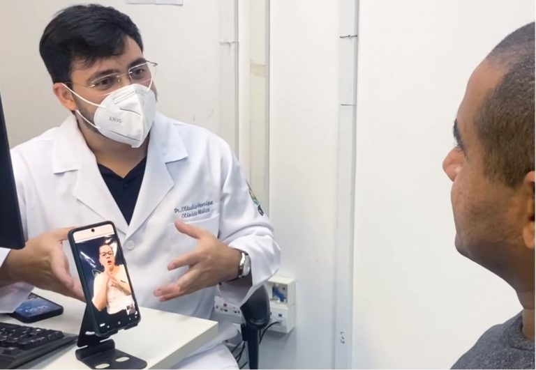 Foto colorida de atendimento em consultório de saúde no Ceará. Na imagem, o médico fala e uma intérprete traduz em Libras em um celular Saúde mental de pessoas surdas é desafio em todo o Brasil
