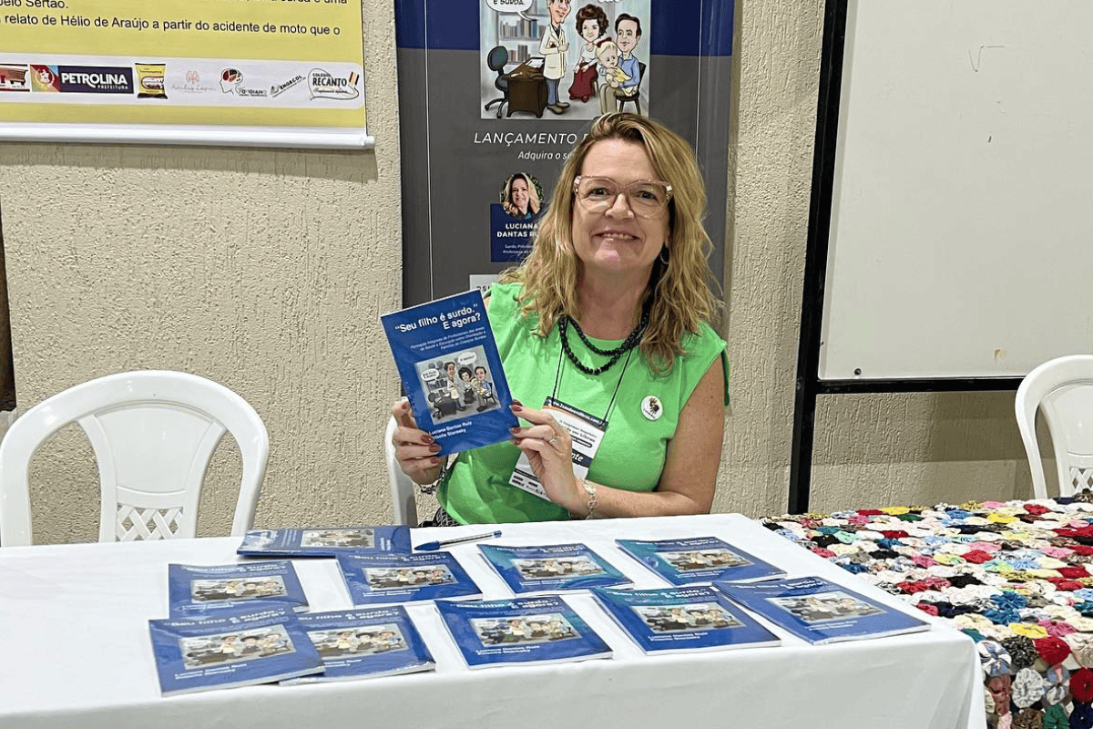 Foto colorida de Luciana Ruiz durante lançamento de livro. Luciana é uma mulher branca e loira. Ela usa uma camisa verde e segura um exemplar do seu livro, ela usa óculos e sorri. Na frente, bancada com outros exemplares do livro.