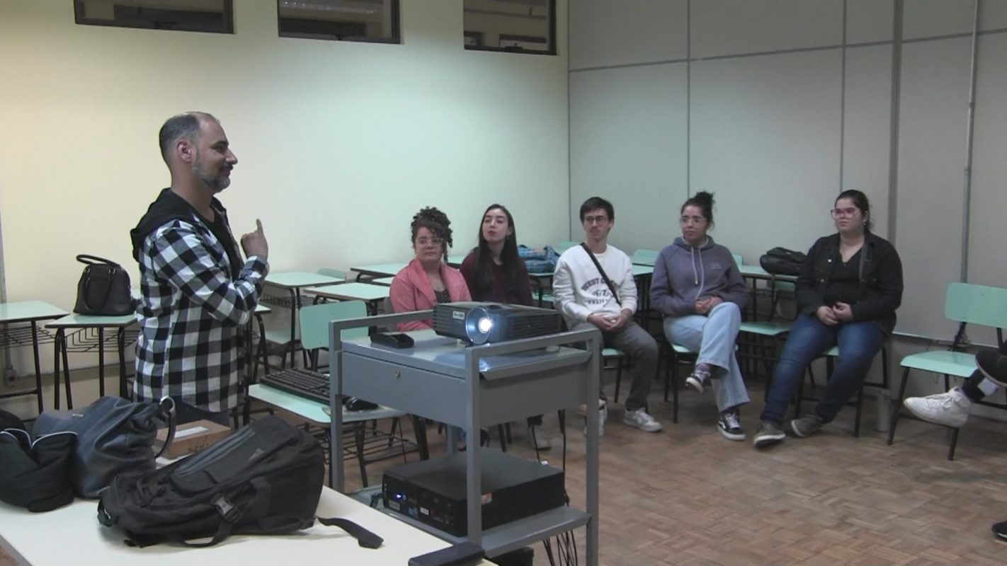 Foto colorida de sala de aula. Willian aparece de pé, fazendo um sinal perto do peito. Ele é um homem branco, com cabelo curto e usa um casaco xadrez. Os alunos aparecem sentados e olham para ele