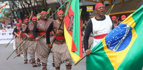 Foto colorida de desfile de lanceiros negros em Caxias do Sul. Em primeiro plano, os lanceiros aparecem vestidos com coletes cinza/dourado e usam bombachas preta e branca e botas de couro. Nos braços empunham lanças; osprimeiros leva uma bandeira do Brasil. Ao fundo, prédios da cidade.