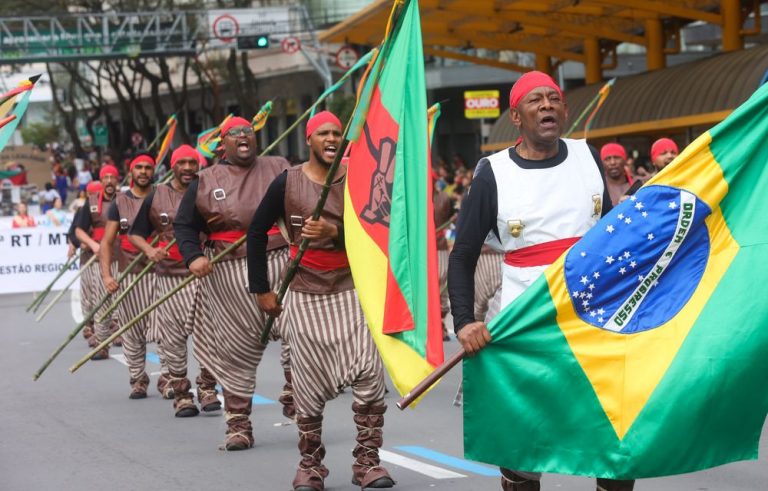 Foto colorida de desfile de lanceiros negros em Caxias do Sul. Em primeiro plano, os lanceiros aparecem vestidos com coletes cinza/dourado e usam bombachas preta e branca e botas de couro. Nos braços empunham lanças; osprimeiros leva uma bandeira do Brasil. Ao fundo, prédios da cidade.