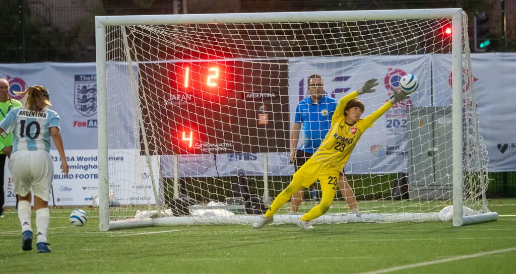 Foto colorida de partida do mundial de futebol de cegas. A imagem mostra um momento decisivo do pênalti cobrado por Yohana Aguilar, jogadora da Argentina. Na foto, Yohana está do lado esquerdo da imagem esperando, e a goleira japonesa está toda esticada para o lado direito pouco antes da bola bater na trave e entrar no gol