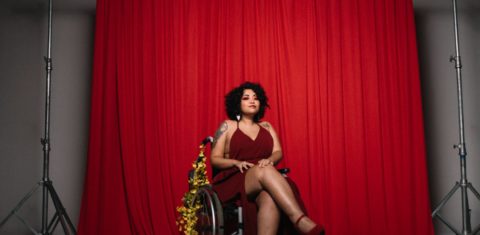 Foto colorida da artista def Amanda Lyra. Ela é uma mulher branca com cabelos cacheados pretos. Ela usa um vestido vermelho e está em uma cadeira de rodas enfeitadas com flores amarelas. Ao fundo, cortina vermelha.