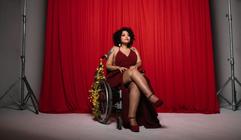 Foto colorida da artista def Amanda Lyra. Ela é uma mulher branca com cabelos cacheados pretos. Ela usa um vestido vermelho e está em uma cadeira de rodas enfeitadas com flores amarelas. Ao fundo, cortina vermelha.
