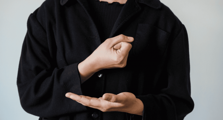 Foto colorida de mãos de um intérprete de Libras. Na imagem, a pessoa aparece com a mão esquerda estendida para a frente do corpo e a mão direita mais acima e fechada. A pessoa usa roupa preta
