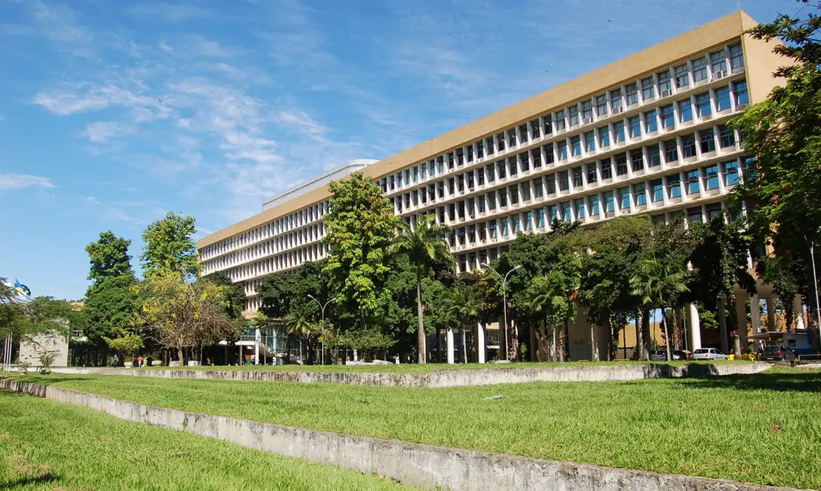 Foto colorida de prédio da UFRJ. Na imagem, aparece o prédio de lado, com diferentes árvores e uma calçada na frente. Ao fundo, céu azul