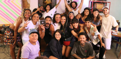 Foto colorida de evento do Slam do Corpo em Ubatuba. Na foto, os participantes do encontro estão reunidos, alguns sentados e outros em pé, eles sorriem e olham para a câmera. Ao fundo, decoração do bar.