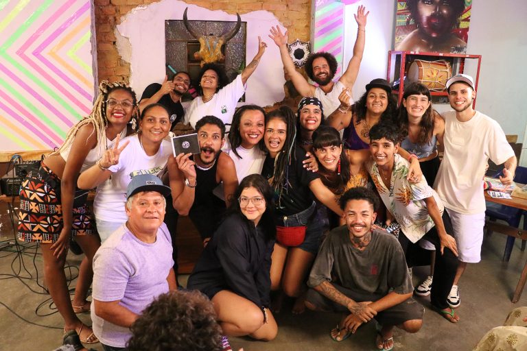 Foto colorida de evento do Slam do Corpo em Ubatuba. Na foto, os participantes do encontro estão reunidos, alguns sentados e outros em pé, eles sorriem e olham para a câmera. Ao fundo, decoração do bar.