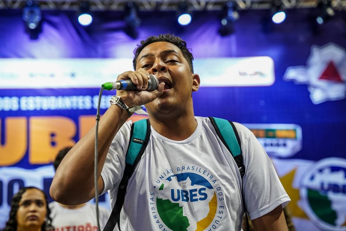 Foto colorida de Hugo Silva. Ele é um jovem preto com cabelo preto. Ele usa uma camisa branca com a logo da ubes e segura um microfone perto da boca