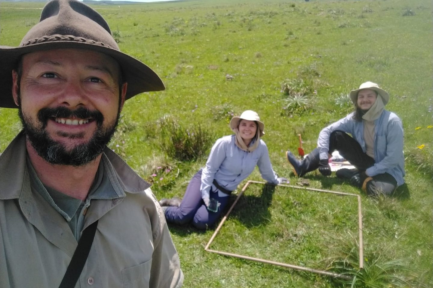 Foto colorida de Fábio, Ana e Mateus junto ao metro quadrado com 64 espécies em campo nativo. Fábio está a esquerda e é um homem branco de barba preta, ele usa um chapéu marrom e sorri. Ao centro, aparece Ana, ela é uma mulher branca e usa um chapéu marrom e blusa azul clara, ao lado dela, sentado no chão Mateus usa um chapéu igual e é um homem branco.