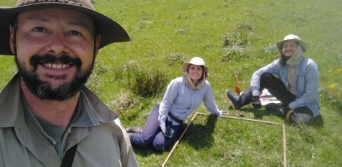 Foto colorida de Fábio, Ana e Mateus junto ao metro quadrado com 64 espécies em campo nativo. Fábio está a esquerda e é um homem branco de barba preta, ele usa um chapéu marrom e sorri. Ao centro, aparece Ana, ela é uma mulher branca e usa um chapéu marrom e blusa azul clara, ao lado dela, sentado no chão Mateus usa um chapéu igual e é um homem branco.