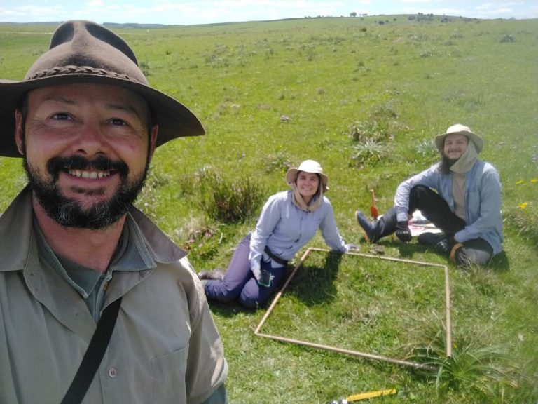 Foto colorida de Fábio, Ana e Mateus junto ao metro quadrado com 64 espécies em campo nativo. Fábio está a esquerda e é um homem branco de barba preta, ele usa um chapéu marrom e sorri. Ao centro, aparece Ana, ela é uma mulher branca e usa um chapéu marrom e blusa azul clara, ao lado dela, sentado no chão Mateus usa um chapéu igual e é um homem branco.