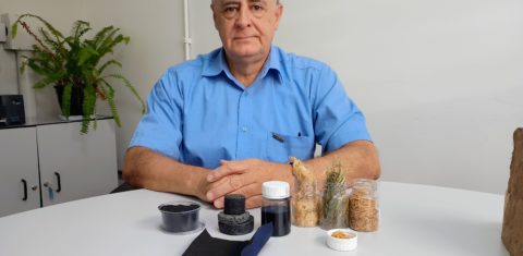 Foto colorida de José Beltran. Ele é um homem branco de cabelos cinzas e usa uma camisa azul. Na frente dele, em uma mesa estão amostras do pó valiente, de casca de arroz e capim-annoni