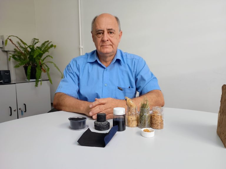 Foto colorida de José Beltran. Ele é um homem branco de cabelos cinzas e usa uma camisa azul. Na frente dele, em uma mesa estão amostras do pó valiente, de casca de arroz e capim-annoni