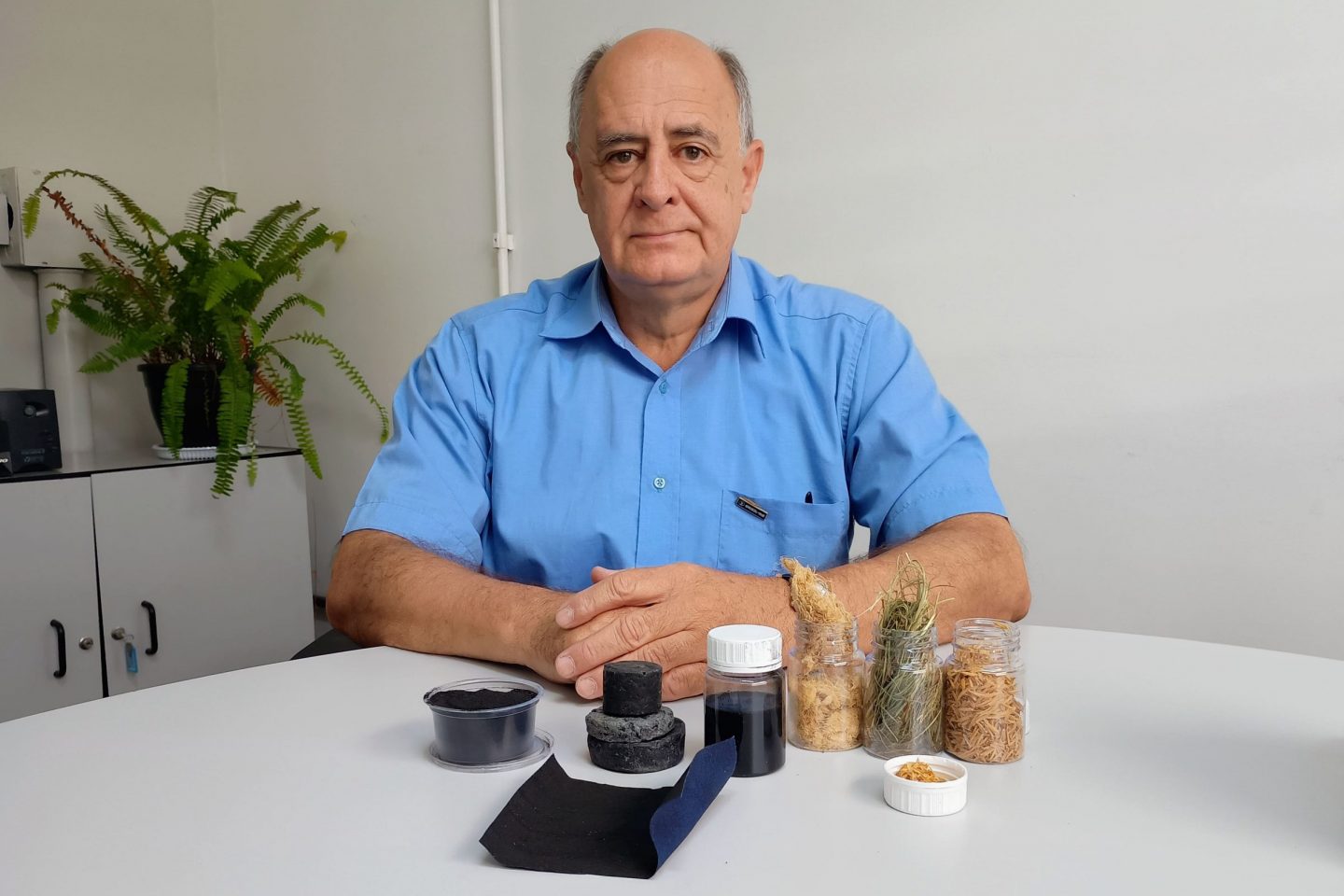 Foto colorida de José Beltran. Ele é um homem branco de cabelos cinzas e usa uma camisa azul. Na frente dele, em uma mesa estão amostras do pó valiente, de casca de arroz e capim-annoni