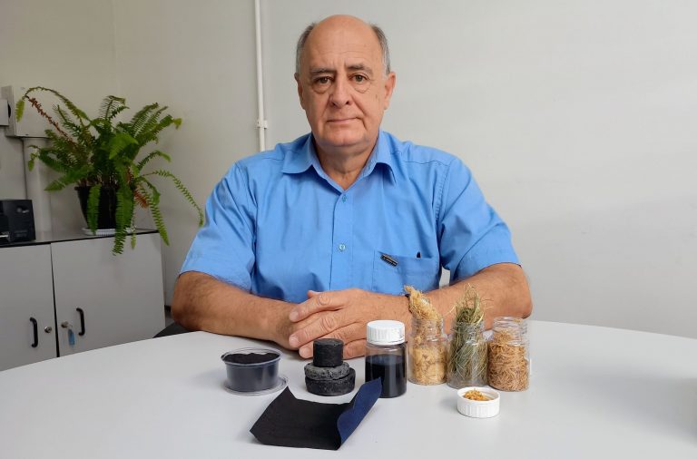 Foto colorida de José Beltran. Ele é um homem branco de cabelos cinzas e usa uma camisa azul. Na frente dele, em uma mesa estão amostras do pó valiente, de casca de arroz e capim-annoni