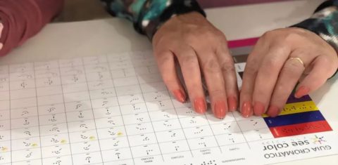 Foto colorida das mãos de uma pessoa sob uma folha com códigos de cores em braille e no See Color