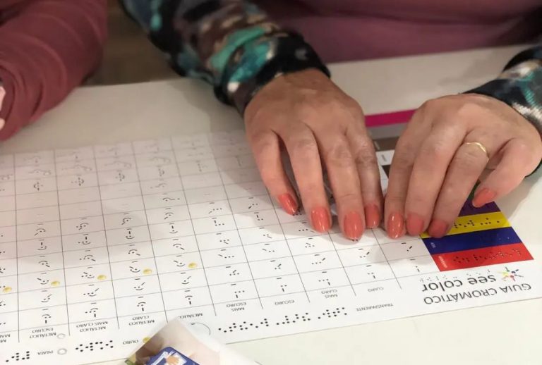 Foto colorida das mãos de uma pessoa sob uma folha com códigos de cores em braille e no See Color
