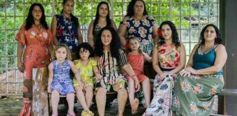 Foto colorida de mulheres ciganas e crianças sentadas e olhando para foto. Elas usam vestidos e saias coloridas, característicos da cultura cigana