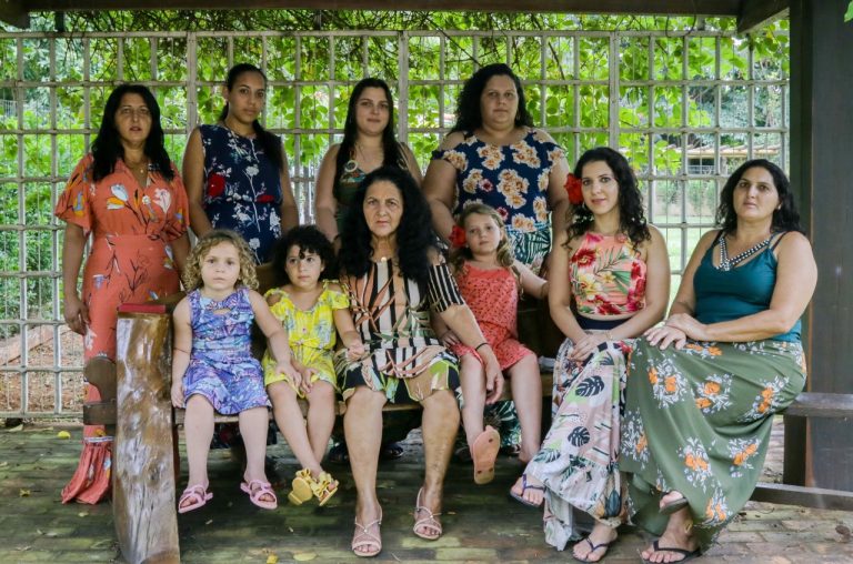 Foto colorida de mulheres ciganas e crianças sentadas e olhando para foto. Elas usam vestidos e saias coloridas, característicos da cultura cigana