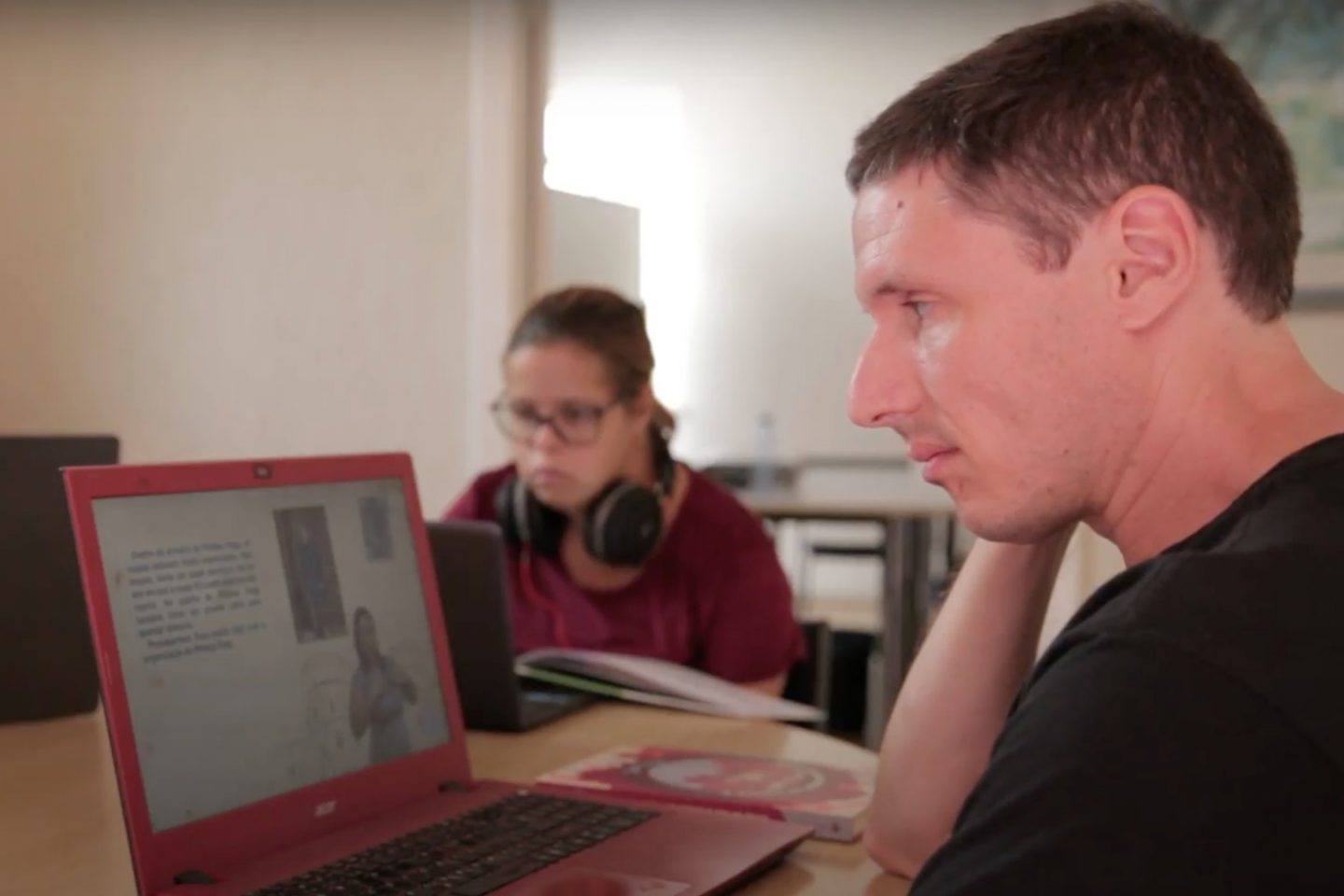 Foto colorida de Mario Paulo Greggio. Mario está à direita da imagem, ele é branco e tem cabelos pretos curtos. Ele usa uma camiseta preta e olha em direção ao computador que está sobre uma mesa no canto esquerdo da imagem. O computador exibe um vídeo de livro acessível. Ao fundo aparece outra pessoa usando um computador.