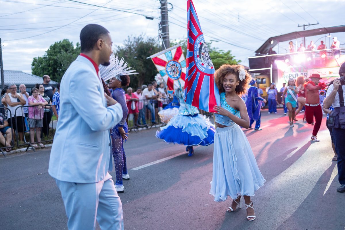 Foto colorida de desfile das escolas de samba de Canoas. Na imagem, dois jovens aparecem em primeiro plano, uma menina com uma bandeira e um menino com um terno que aparece de costas e olhando para ela