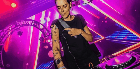 Foto colorida da DJ surda Lenny Vibe. Lenny é uma mulher branca, com cabelo preto e ela usa uma camiseta preta. Ela possui tatuagens no braço direito, que está estendido em direção a um mixer. Ao fundo, luzes de festa