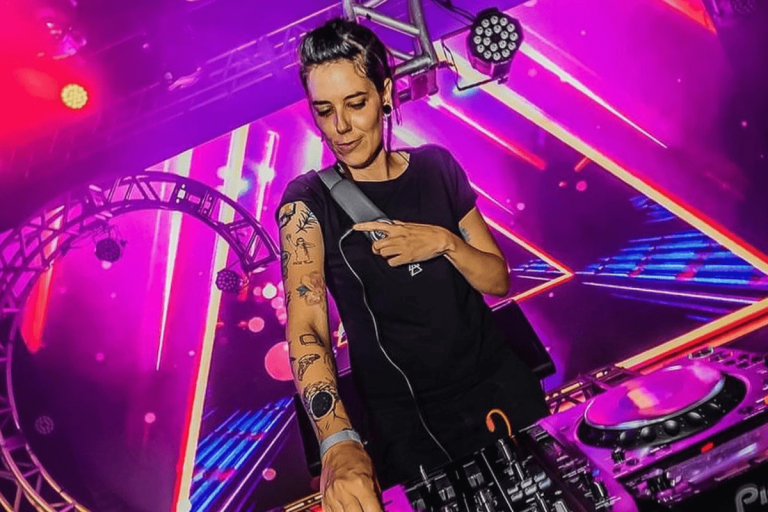 Foto colorida da DJ surda Lenny Vibe. Lenny é uma mulher branca, com cabelo preto e ela usa uma camiseta preta. Ela possui tatuagens no braço direito, que está estendido em direção a um mixer. Ao fundo, luzes de festa