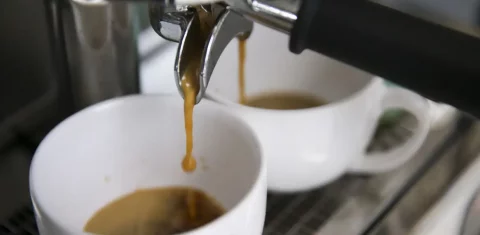Foto colorida de xícara de café sendo enchida. Na imagem, parte da bebida aparece nas xícaras brancas e parte caindo de torneira