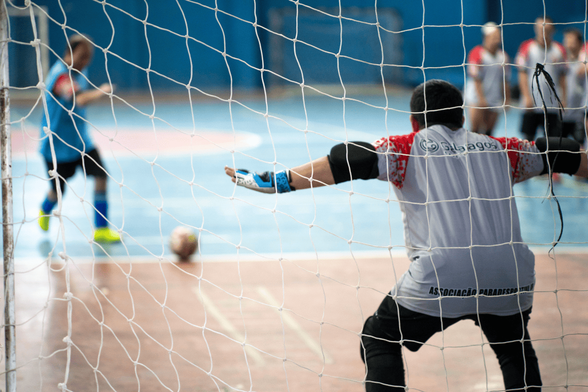 Foto colorida de treino de futsal down. Em primeiro plano, o goleiro aparece de costas e braços abertos. Ao fundo, outro atleta domina a bola