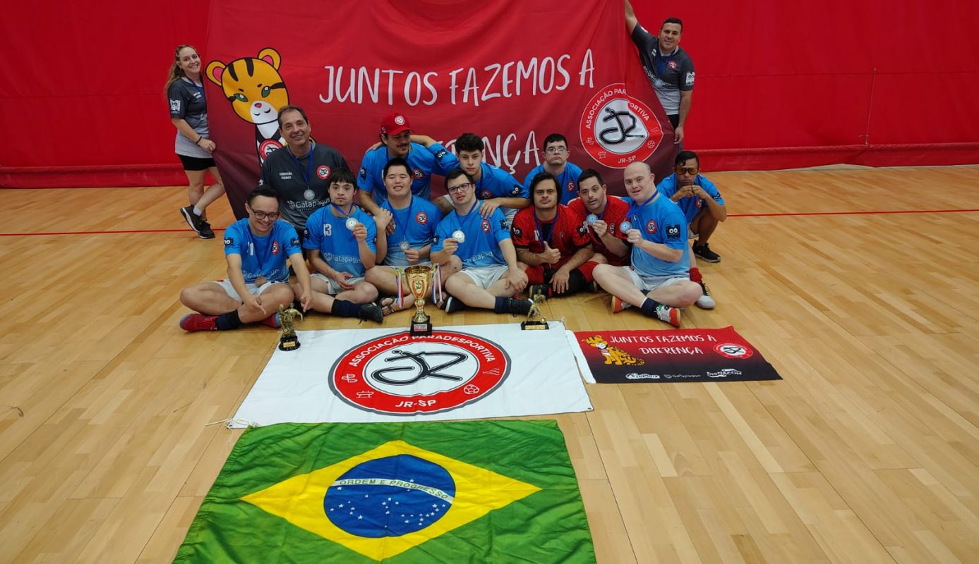 Foto colorida de atletas da Associação JR sentados diante da bandeira do projeto e do Brasil. Eles usam camisetas azuis e estão sorrindo. Na frente deles está uma taça de campeão