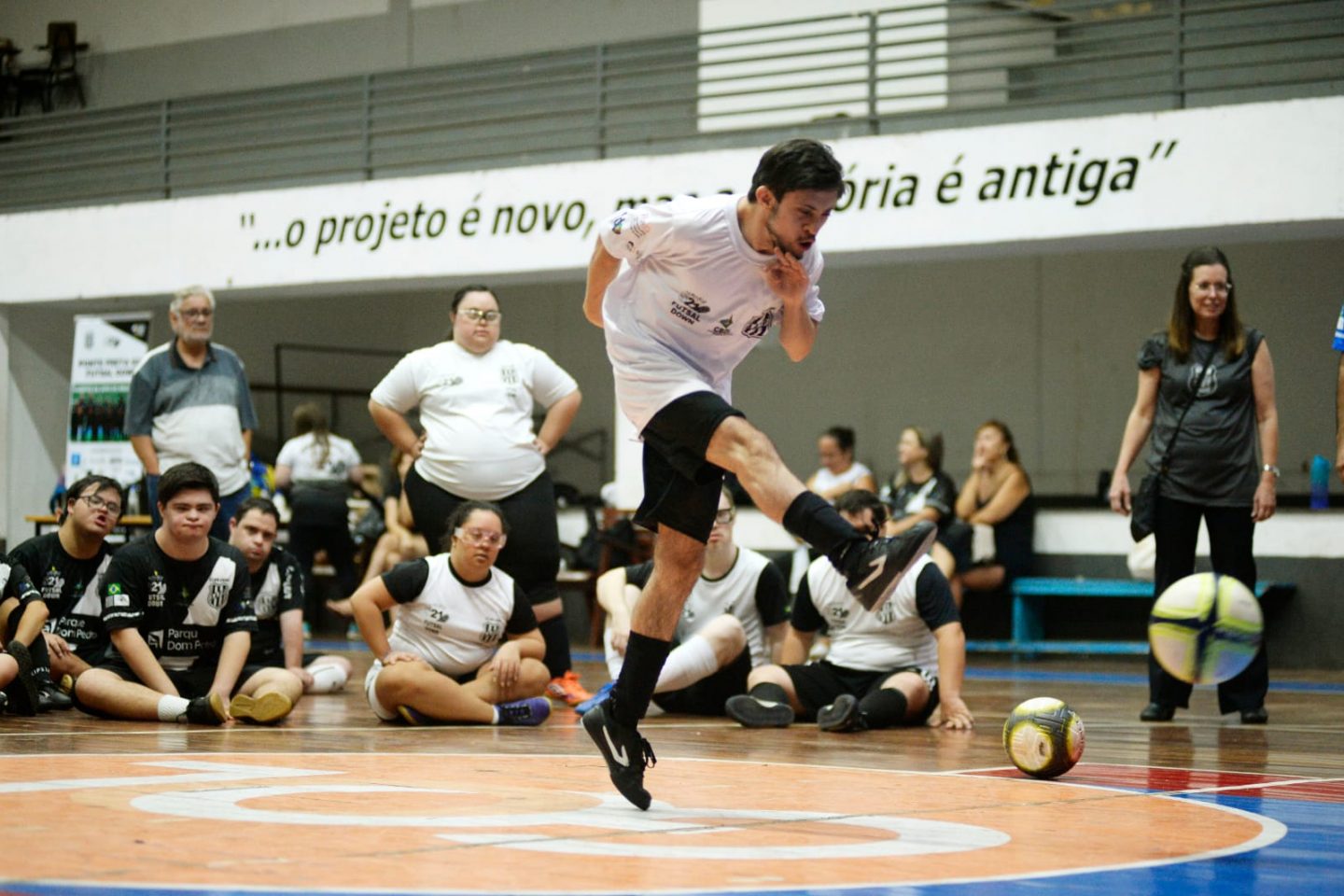 Foto colorida de atleta de futsal down. Na imagem, o jogador aparece no ar, após realizar um chute. Ao fundo, outros atletas aparecem sentados