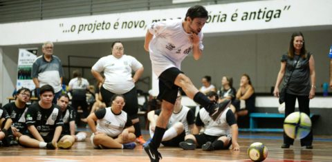 Foto colorida de atleta de futsal down. Na imagem, o jogador aparece no ar, após realizar um chute. Ao fundo, outros atletas aparecem sentados