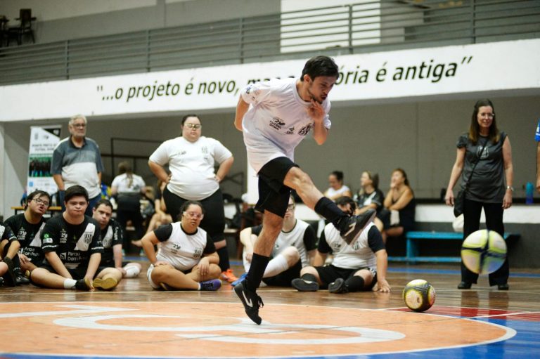 Foto colorida de atleta de futsal down. Na imagem, o jogador aparece no ar, após realizar um chute. Ao fundo, outros atletas aparecem sentados
