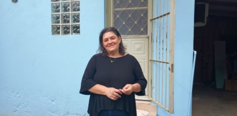 Foto colorida de Suzi Bolsan Post. Ela é uma mulher branca com cabelos pretos curtos. Ela usa uma blusa preta e sorri, com os braços juntos em frente ao corpo. Ao fundo, porta e parede de sua casa em Santa Maria.