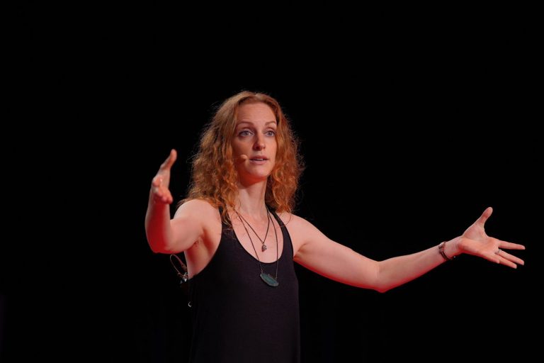 Foto colorida de Laila Zaid em pé durante evento do TED. Laila é uma mulher branca, com cabelos ruivos. Ela está com uma camiseta preta e com os braços abertos. Ao fundo, parede preta.