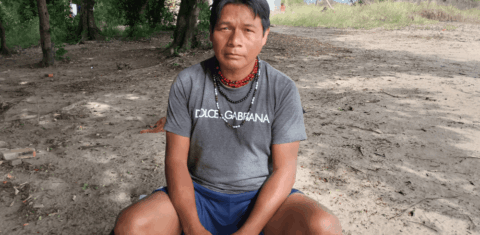 Foto colorida de Cesário Timóteo. Ele é um Guarani com cabelo preto e sem barba. Ele está sentado e usa uma camiseta cinza e vários colares. Ao fundo, aparece terreno, árvores e uma casa