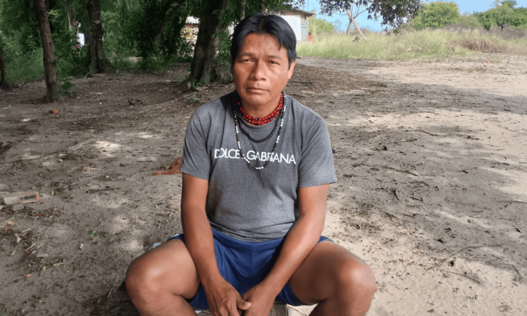 Foto colorida de Cesário Timóteo. Ele é um Guarani com cabelo preto e sem barba. Ele está sentado e usa uma camiseta cinza e vários colares. Ao fundo, aparece terreno, árvores e uma casa