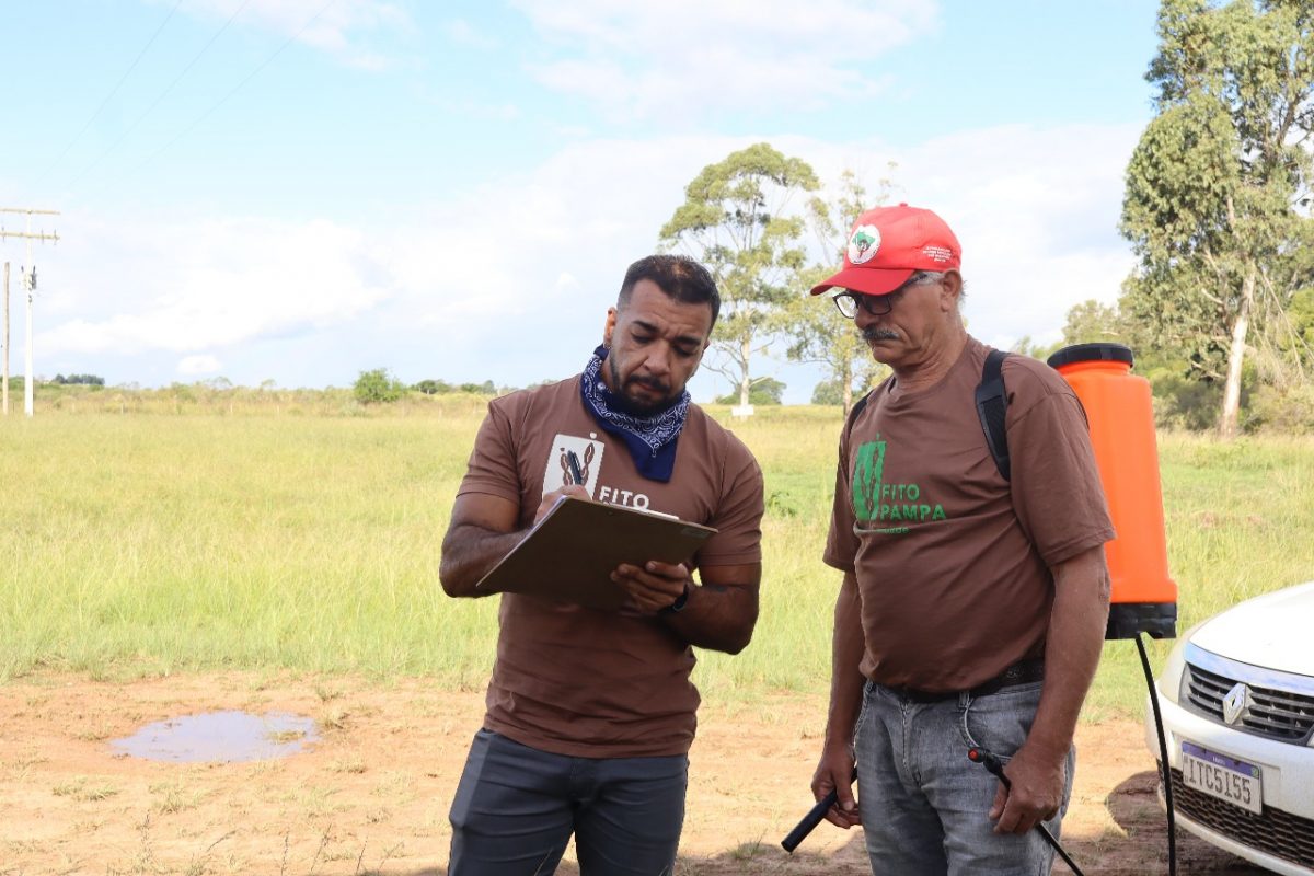 Foto colorida de dois membros do FitoPampa dialogando. Um dos homens segura uma prancheta e o outro está com um tanque para passar produtos nas costas.