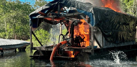 Foto colorida de destruição de estrutura do garimpo ilegal na Terra Indígena Kayapi (MT); mineração ilegal na Amazônia. Na imagem, aparece um galpão em chamas na beira de um rio.