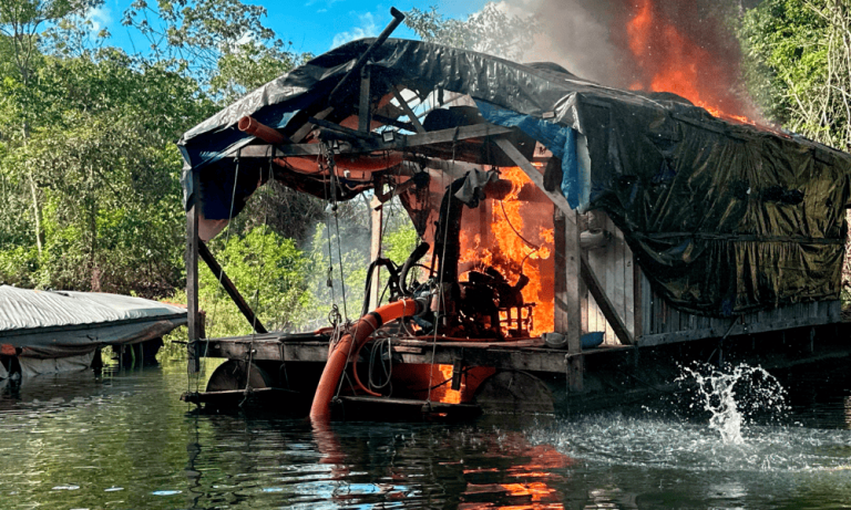 Foto colorida de destruição de estrutura do garimpo ilegal na Terra Indígena Kayapi (MT); mineração ilegal na Amazônia. Na imagem, aparece um galpão em chamas na beira de um rio.