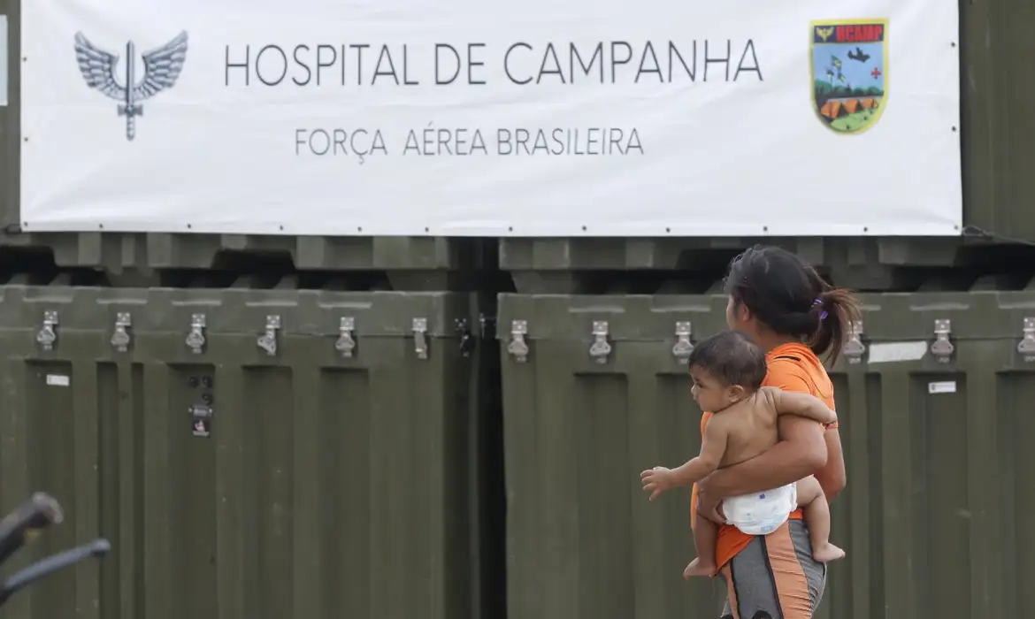 Foto colorida de uma mulher com uma criança no colo de costas, diante do Hospital de Campanha da Força Aérea Brasileira em Roraíma
