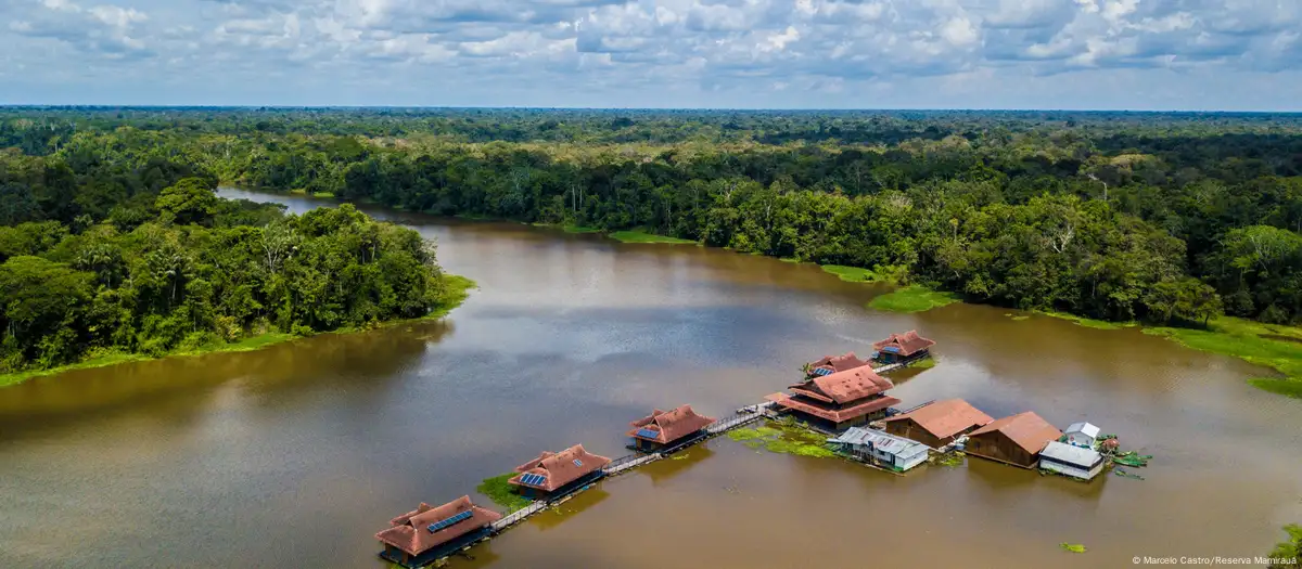 Foto colorida da Reserva de Desenvolvimento Sustentável Mamirauá. Na imagem, aparece um rio com diferentes casas suspensas e um floresta ao fundo.
