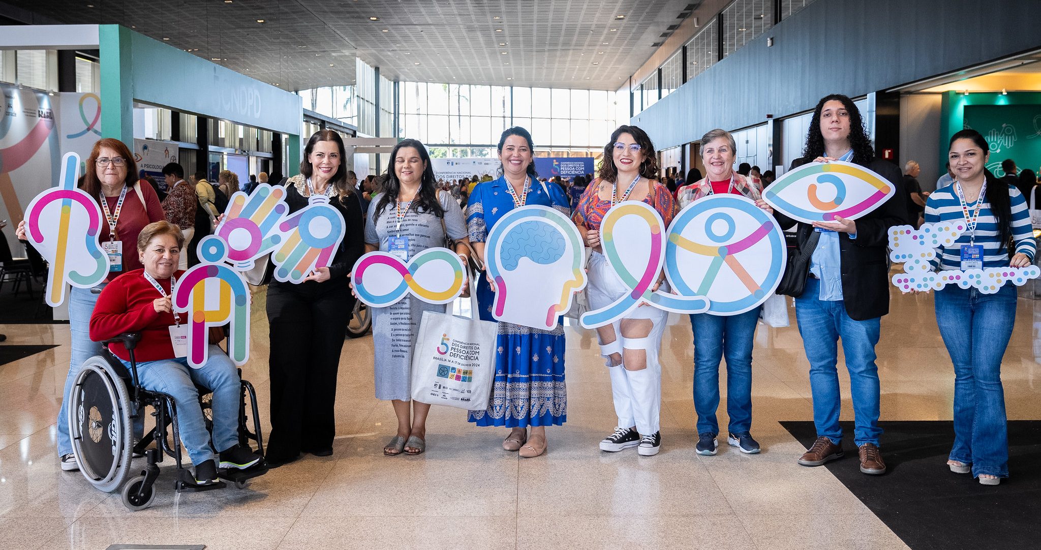 Foto colorida de pessoas reunidas durante a 5°Conferência Nacional dos Direitos das Pessoas com Deficiência. Na imagem, as pessoas seguram figuras que representam diferentes deficiências
