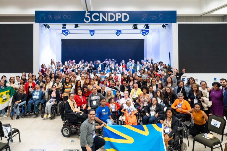 Foto colorida de um dos encontros da 5ª Conferência Nacional dos Direitos das Pessoas com Deficiência. Na imagem, aparecem diversas pessoas com deficiência reunidas na frente do palco, em Brasília (DF)