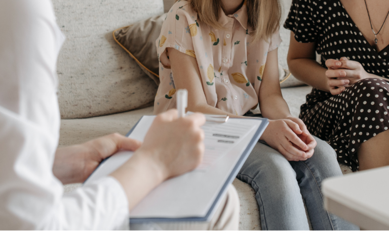 Foto colorida de criança ao lado da mãe em consulta médica. Na imagem, aparece um prontuário em primeiro plano e uma menina ao lado de uma mulher ao fundo. Discussão sobre direitos de mães trabalhadoras envolve acompanhamento de crianças em consultas
