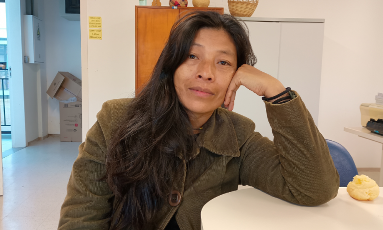 Foto colorida de Patrícia Ferreira Ixapy. Ela é uma mulher indígena guarani de cabelos pretos, lisos e longos. Ela usa um casaco marrom e está com o braço esquerdo apoiado em uma mesa. Ao fundo, armário e parede branca