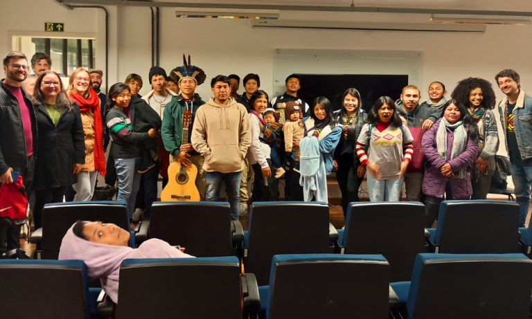 Foto colorida de estudantes Mbyá-Guarani e o público no auditório do prédio 40A da UFSM, em Santa Maria. Na imagem, as pessoas aparecem em grupo posando para a foto.